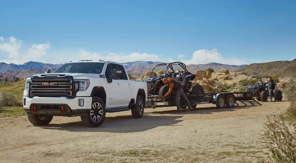 A white 2022 GMC Sierra 2500 AT4 is shown from the front at an angle while hooked up to a trailer carrying UTVs.