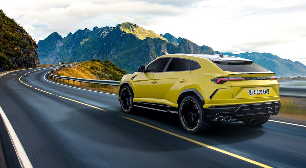 A yellow 2020 Lamborghini Urus is shown driving past a lake in the mountains.