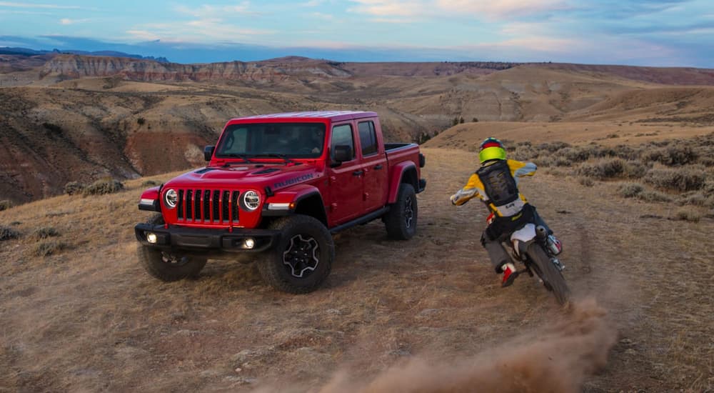 A red 2020 Jeep Gladiator Rubicon is shown from the front at an angle with a dirt-bike riding by.