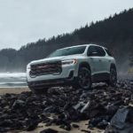 A white 2023 GMC Acadia AT4 is shown from the front on a rocky shore.