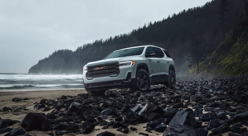 A white 2023 GMC Acadia AT4 is shown from the front on a rocky shore.