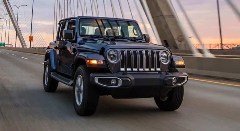 A grey 2022 Jeep Wrangler is shown from the front at an angle while driving over a bridge after leaving a used Jeep dealer.