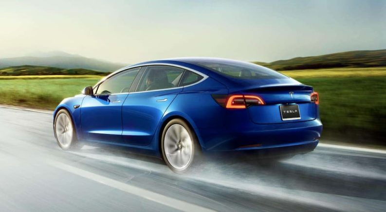 A blue 2020 Tesla Model 3 is shown from a rear angle driving past a field.