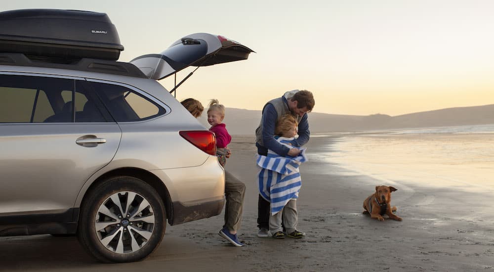 A family and a dog are shown next to a silver 2015 Subaru Outback at a beach after researching how to sell their car.