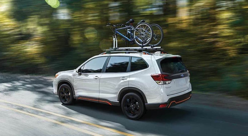 A white 2021 Subaru Forester Sport is shown from the side driving on a wood-lined road.