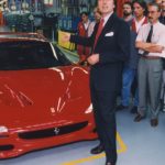 A group of people are shown standing around a red 1995 Ferrari F50.