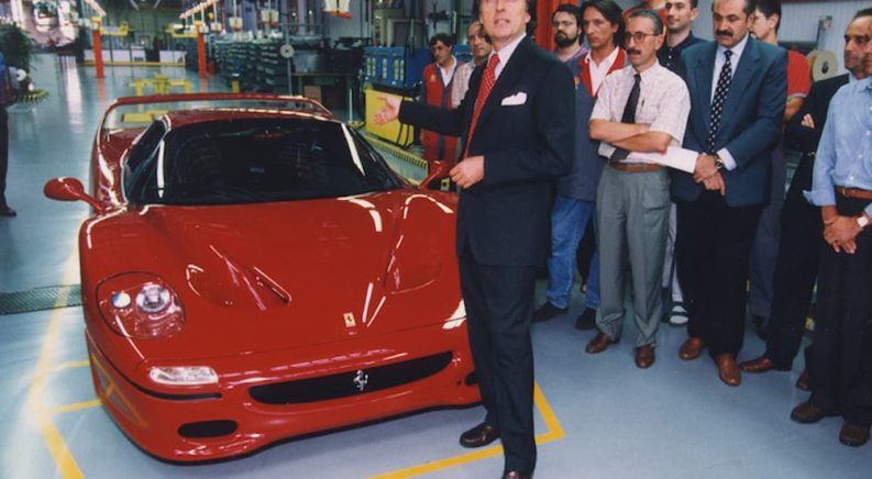 A group of people are shown standing around a red 1995 Ferrari F50.