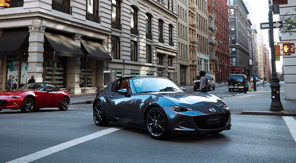 A grey 2022 Mazda MX-5 Miata RF is shown driving through an intersection after leaving a Mazda dealer. 