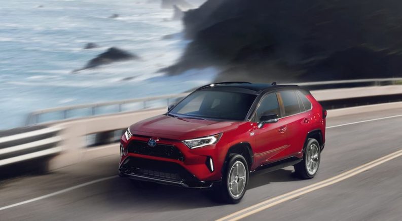 A red 2021 Toyota RAV4 Prime is shown driving on a highway next to the ocean.