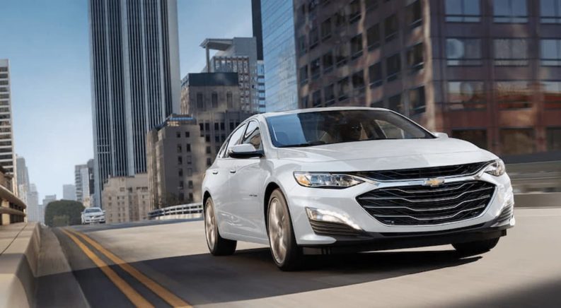 A white 2023 Chevy Malibu is shown driving on a city highway.
