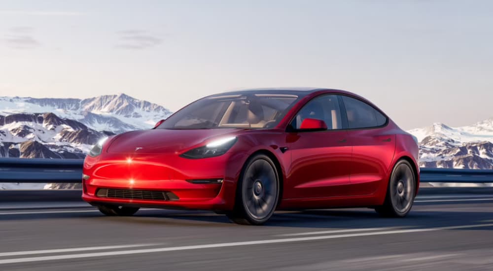 A red 2021 Tesla Model 3 is shown driving on an open highway.