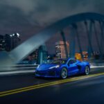 A blue 2024 Chevy Corvette E-Ray 3LZ is shown driving over a city bridge at night.