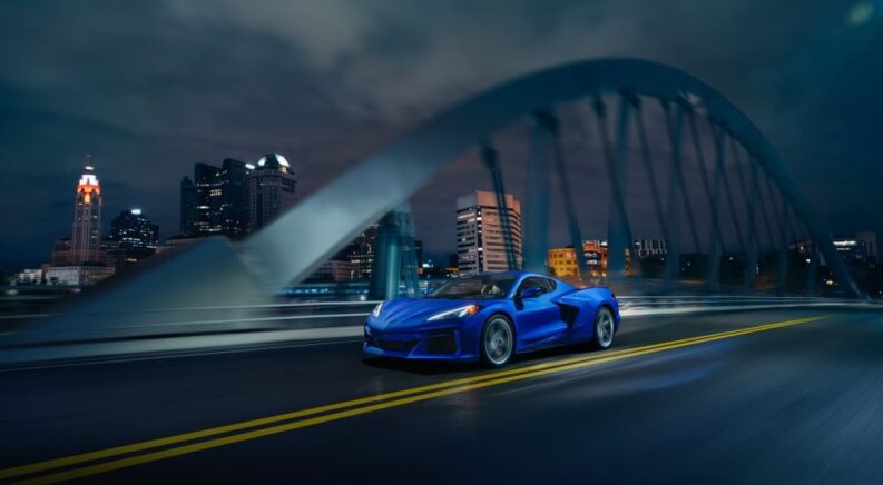 A blue 2024 Chevy Corvette E-Ray 3LZ is shown driving over a city bridge at night.