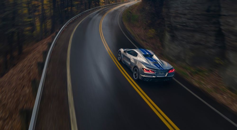 A silver 2024 Chevy Corvette E-Ray 3LZ is shown from the rear driving on a tree-lined road.