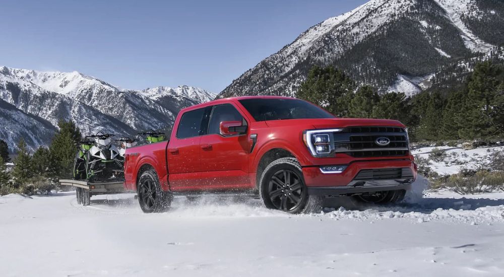 A red 2023 Ford F-150 for sale is shown towing four snowmobiles on a flatbed in the snow.