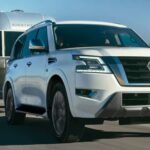 A white 2023 Nissan Armada for sale is shown towing a camper down an open road.