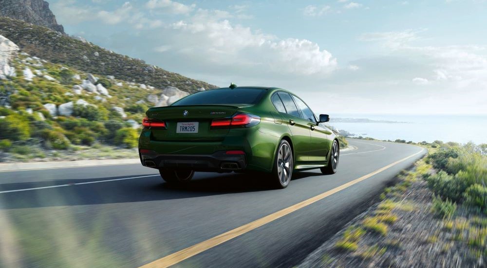 A green 2023 BMW M550i is shown from the rear driving on a coastal road.