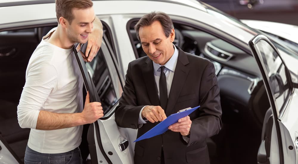A buyer is shown working with a car salesman in front of a white SUV at a used car dealer.