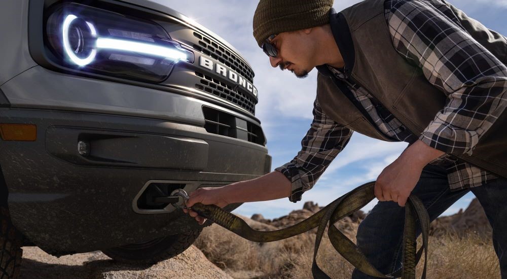 A man is shown attaching a strap to the tow hook of a silver 2023 Ford Bronco Sport Badlands.