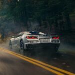 A silver 2024 Chevy Corvette E-Ray with blue racing stripes is shown speeding down a country road.