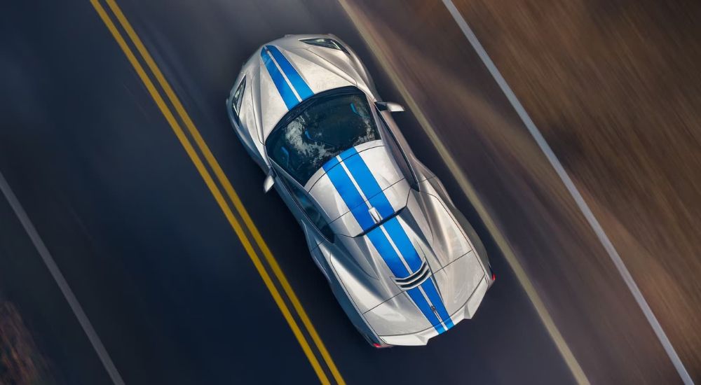 A silver Corvette E-Ray with blue racing stripes is shown from above speeding down a highway. 