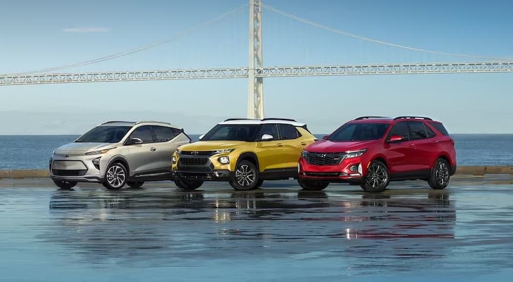 A silver 2023 Chevy Bolt EUV, yellow 2023 Chevy Trailblazer, and red 2023 Chevy Equinox are shown lined up at a Chevy dealer next to the ocean with a suspension bridge in the background.