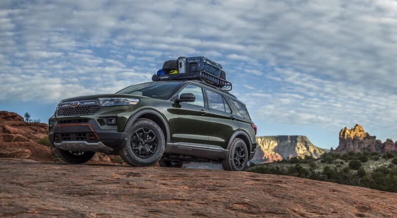 A green 2023 Ford Explorer Timberline loaded with camping gear on its roof rack is shown parked on a rocky outcropping with mountains in the background.