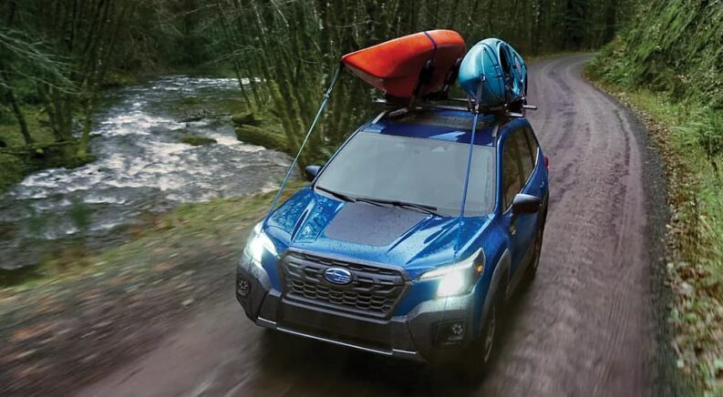 A blue 2023 Subaru Forester Wilderness is shown driving on a dirt road near a stream.