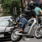 A person fitted in some of the best motorcycle gear is shown driving a grey SONDORS Metacycle through a city street.