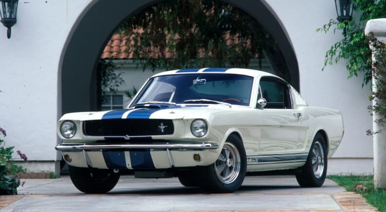 A white and blue 1965 Ford Mustang Shelby GT350 is shown parked on a driveway.