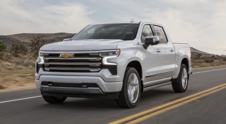 A white 2024 Chevy Silverado 1500 High Country is shown driving on a highway after visiting a Chevy dealer.