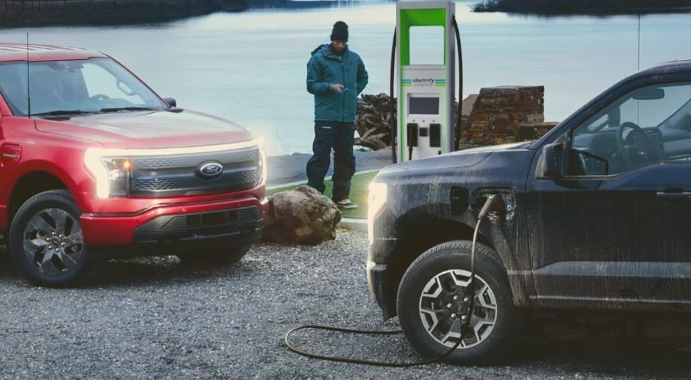 A red and a black 2023 Ford F-150 Lightning are shown charging at a lake.