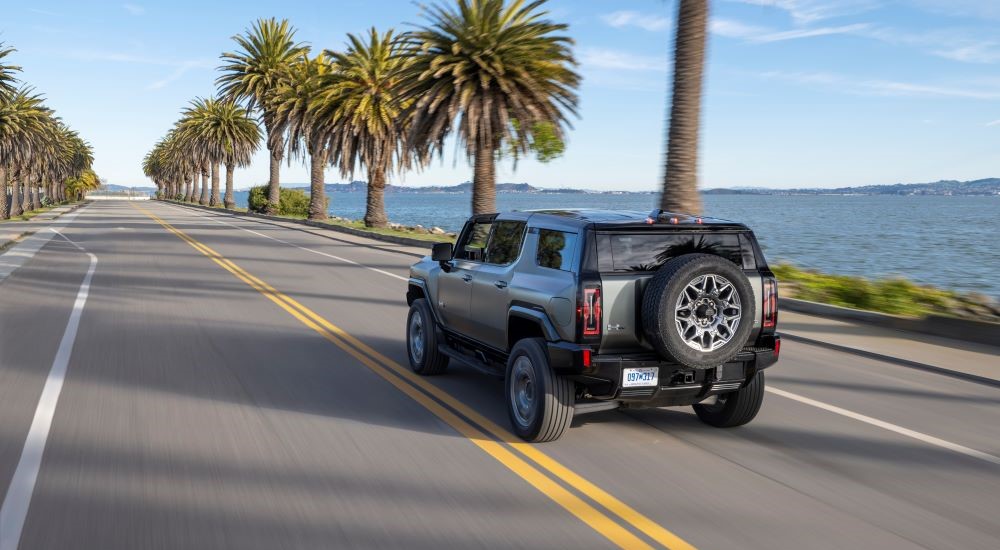 A light green 2024 GMC Hummer EV is shown driving past palm trees.