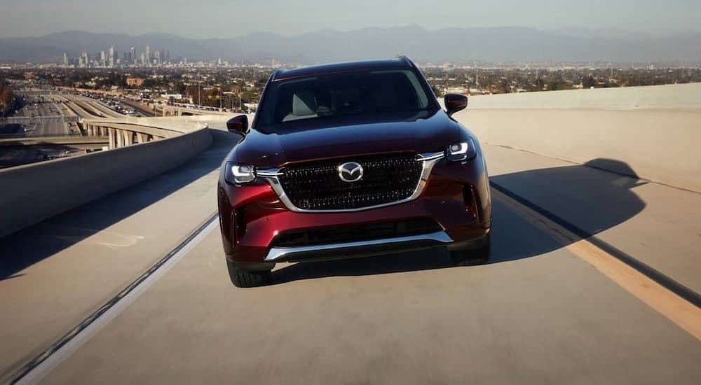 A burgundy 2024 Mazda CX-90 is shown driving on an on-ramp. 