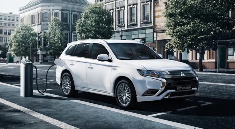 One of the more common electric vehicles for sale, a white 2022 Mitsubishi Outlander PHEV, is shown parked and charging on a city street.