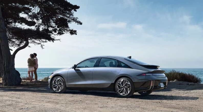 A model of Hyundai IONIQ for sale, a grey 2023 Hyundai IONIQ 6 is shown parked on a beach, with a couple looking out over the sea.