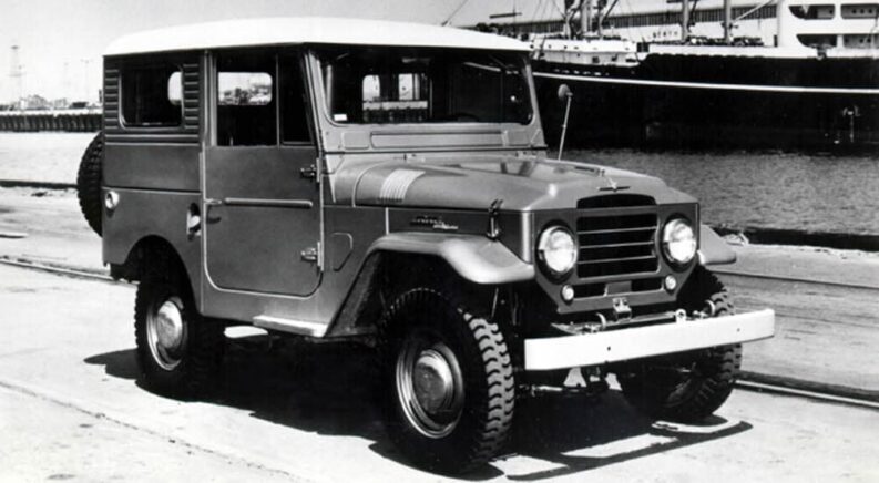 A black-and-white photograph of a 1960 Toyota Land Cruiser FJ40 driving from a used car dealer is shown.
