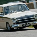 A white 1966 Lotus Cortina is shown driving from a Ford dealer.