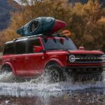 Available at Ford dealerships today, a red 2024 Ford Bronco Outer Banks is shown driving through a shallow pond.