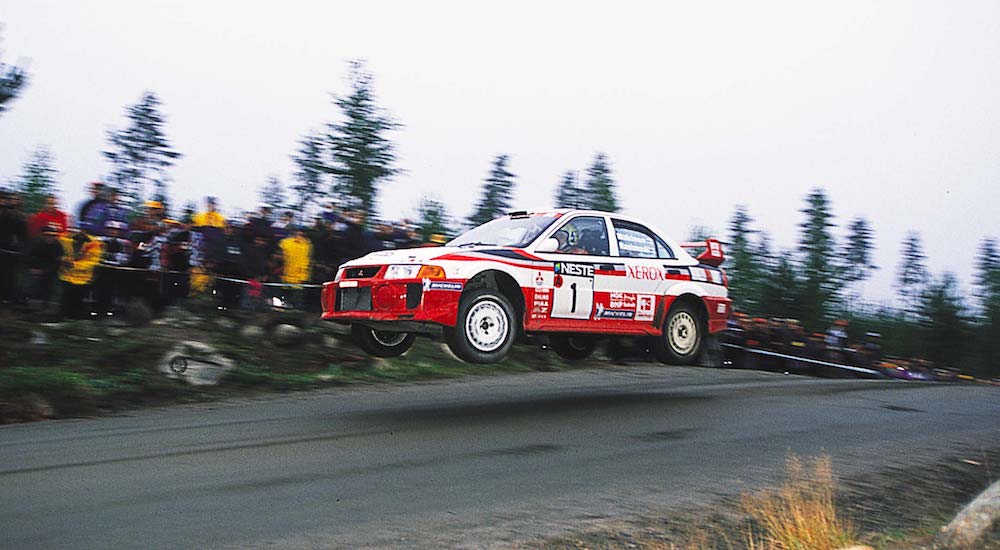 A red-and-white 1992 Mitsubishi Lancer Evolution is shown racing at a rally.