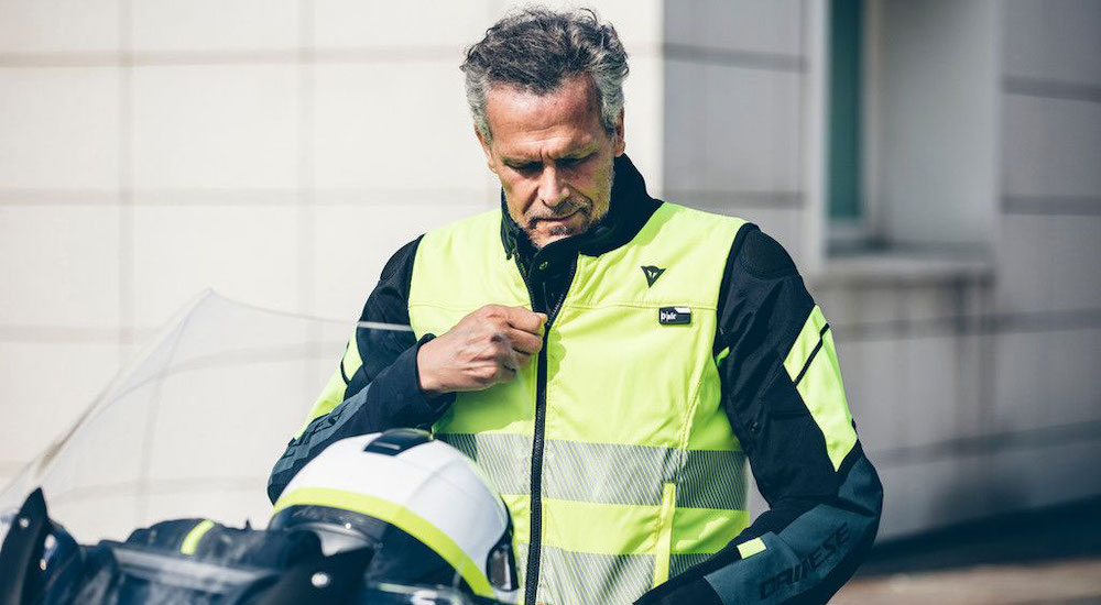 A man sitting on a motorcycle and wearing a Dainese Smart Jacket Hi Vi vest is shown.