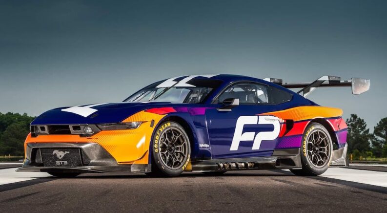 An orange and blue 2024 Ford Mustang GT3 is shown on a race track.