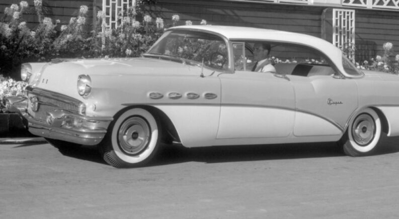 A black-and-white photo of a 1956 Buick Super Riviera parked near a used Buick dealer is shown.