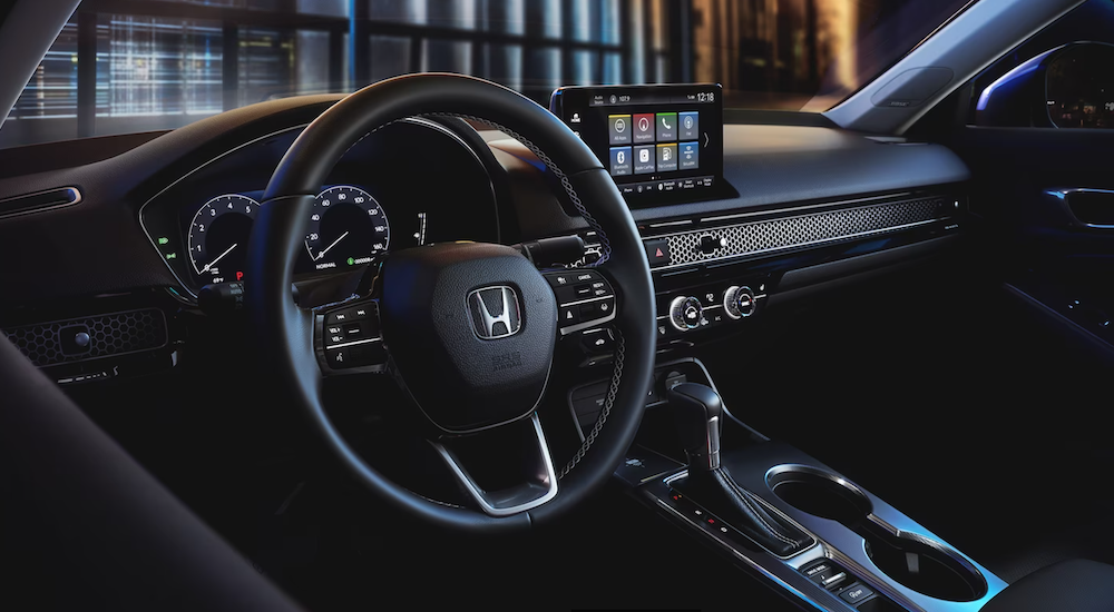 The black interior of a 2024 Honda Civic is shown as seen from the driver's seat, including the steering wheel and dashboard screen.