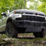A variant Chevy Silverado 1500 for sale, a white 2023 Chevy Silverado ZR2 Bison, is shown parked off-road in a forest.