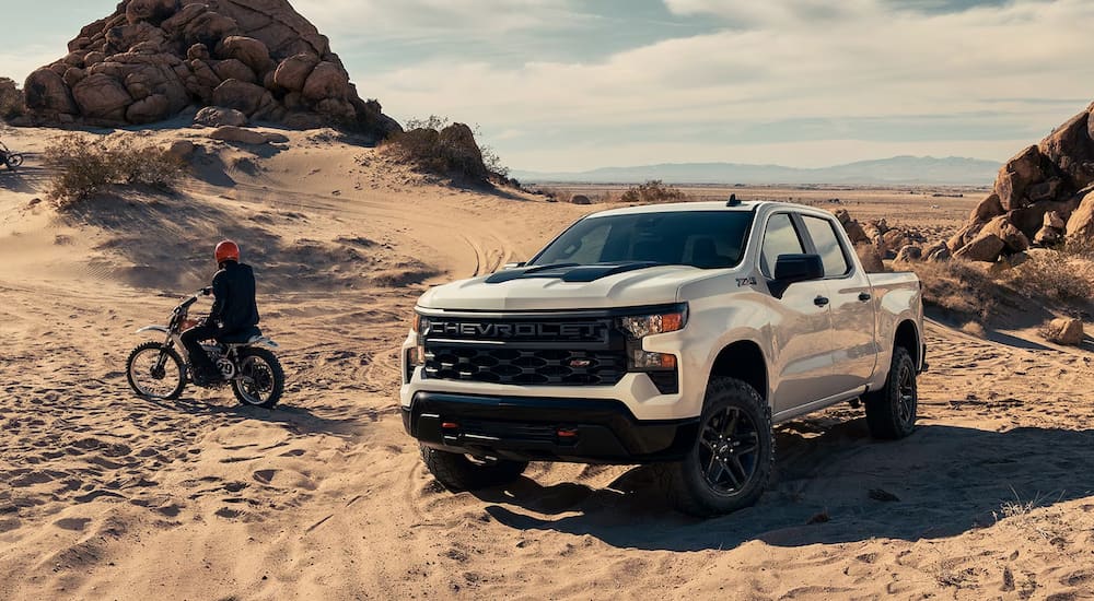 A white 2024 Chevy Silverado 1500 Custom Trail Boss off-roading in dunes with dirt bikes.