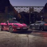 A pink 2024 Porsche 718 Boxter and grey 2024 Porsche 718 Cayman parked at an industrial site.