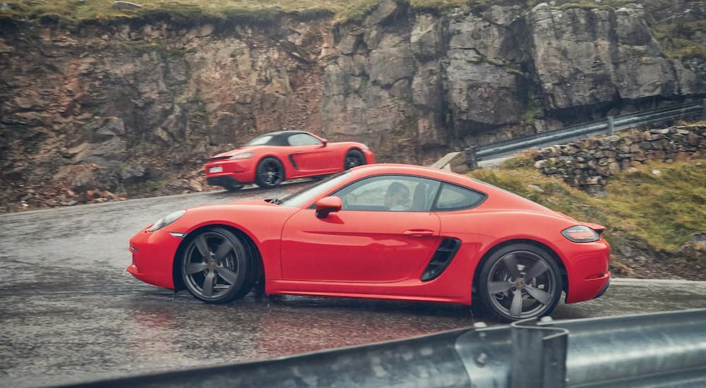 A red 2024 Porsche 718 Boxter and a red 2024 Porsche 718 Cayman driving on a switchback near cliffs.
