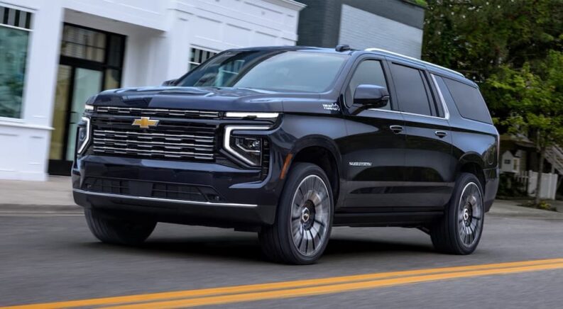A black 2025 Chevy Suburban High Country is shown driving from a dealer with a Chevy for sale.