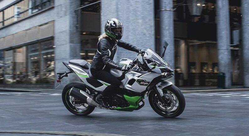 A silver and green Kawasaki Ninja 7 HEV is shown riding near a store.